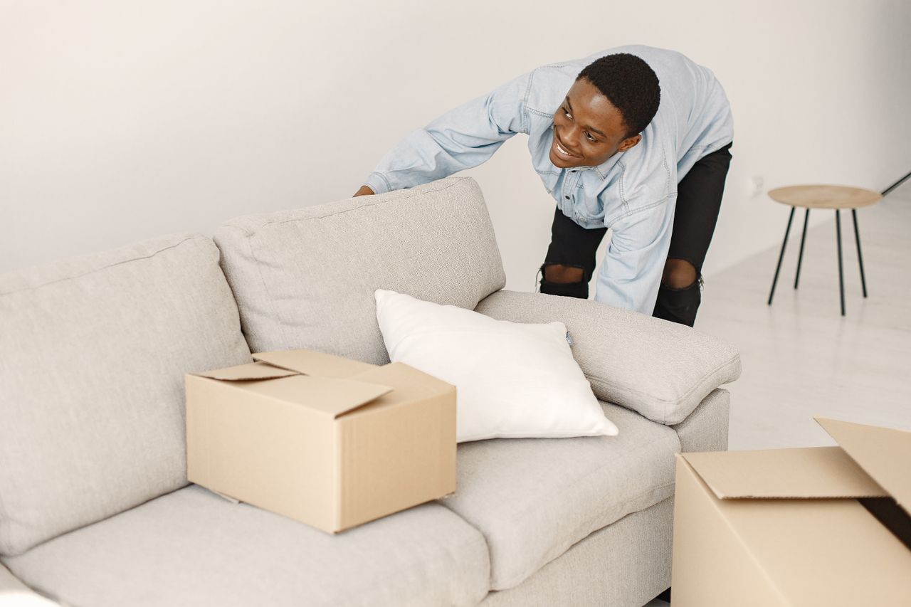 hero-img Young man smiling while arranging cardboard boxes in a modern living room.